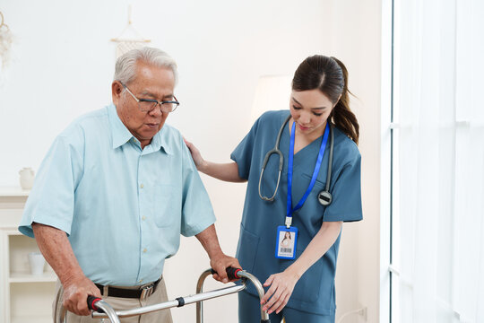Young friendly female caregiver nurse in blue scrubs is smiling and helping Asian senior man practice walking with walker in cozy living room at nursing home. Physical therapy, healthcare concept