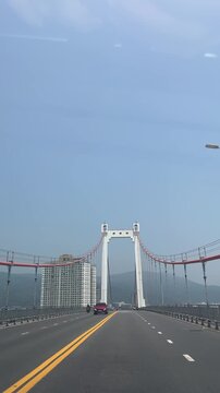 A modern suspension bridge spans a wide road under a clear blue sky. The bridge features white arches and red cables, with mountains visible in the background.