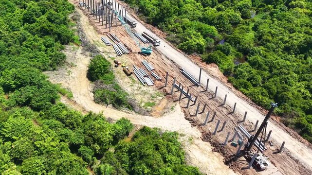 High-angle aerial drone video moving forward over a bridge construction site featuring vertical concrete piles, pile driving equipment, and heavy machinery during infrastructure development.