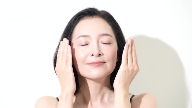 [スローモーション]美しい肌の50代女性のビューティーポートレート、カメラ目線の笑顔　頬に手で触れるスキンケアとエイジングケアイメージの動画
