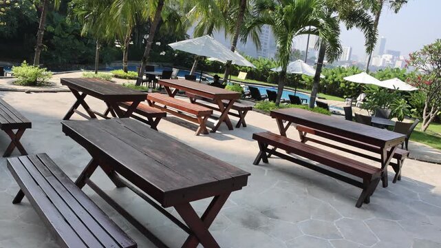 Sunlit patio holds rustic wooden tables and benches. Tropical palms sway gently in the breeze. White umbrellas shade the open-air seating area. Calm pool water reflects the bright sky