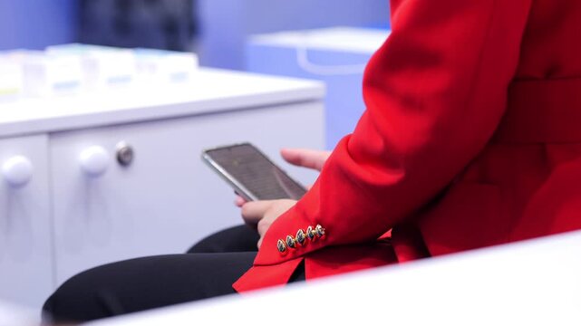 Person in red jacket uses smartphone at event. White furniture and blue background visible. Focus on hands and device interaction. Modern indoor setting with clean lines