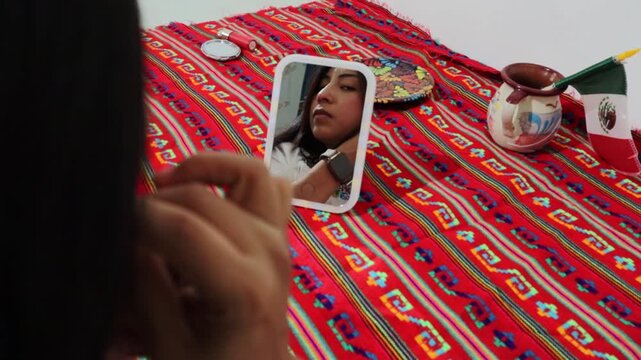 Mexican woman putting on traditional earrings while looking in the mirror on a colorful tablecloth with a small mexican flag, showcasing national pride and indigenous culture