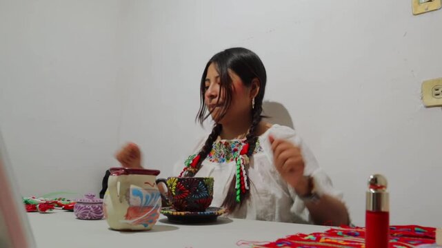 Expressive mexican woman wearing a traditional blouse with braids dancing happily while sitting at a table with colorful artisan crafts and listening to music