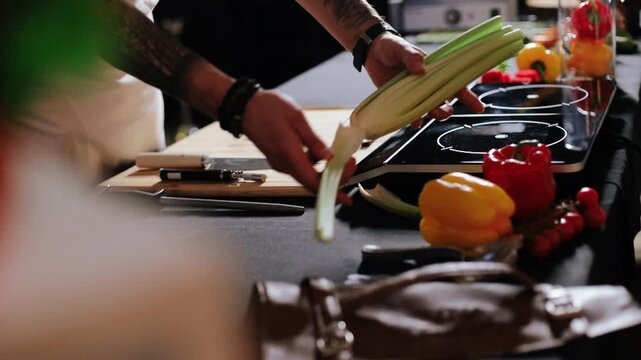 Low-angle close-up of adult hands and forearms separating and arranging celery stalks on wooden cutting board beside glossy induction cooktop, one hand stabilizes bunch while other pulls or places