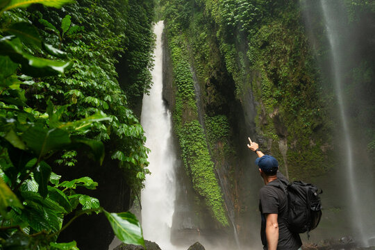 A hiker points at a large waterfall surrounded by lush greenery