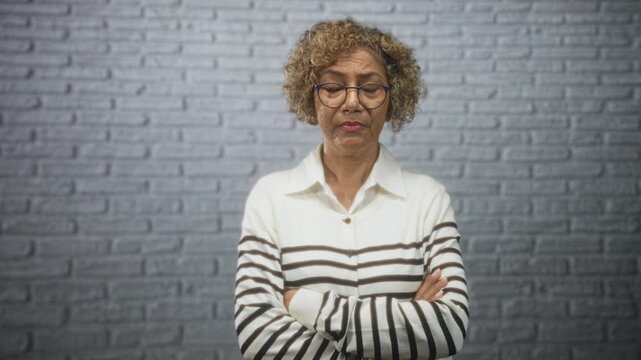 Woman wearing glasses and collared white striped sweater, arms crossed, standing against a brick wall in a studio; quiet concern.