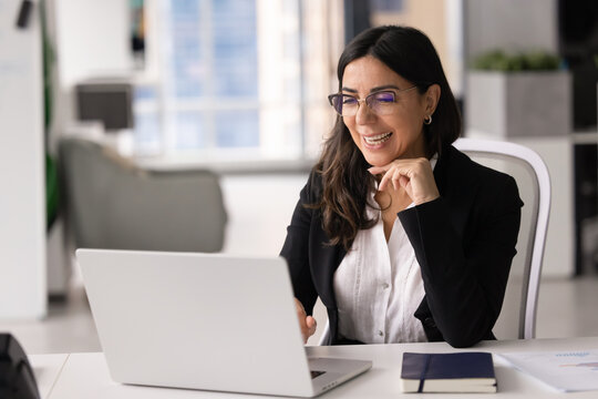 Middle-age businesswoman sits office desk, working on business project on laptop, engage in client consultation, performs online tasks, reviewing positive work results, smiling, achieve corporate goal