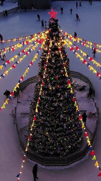 Festive evening at an outdoor ice rink with people skating around a large, illuminated christmas tree adorned with colorful garlands and a bright red star, enjoying winter holidays