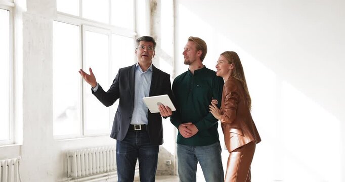 Real estate broker or interior designer showing digital visualization of dream home to married couple on tablet in empty room shaking hands with clients as they hug celebrating final housing decision