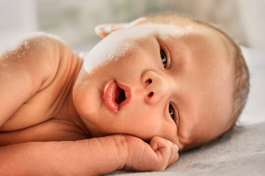 An awake newborn baby lies on pale bedding with an open mouth and a tiny hand beside the cheek. Warm light reveals delicate skin and fine hair in an intimate portrait of the infant gazing forward.