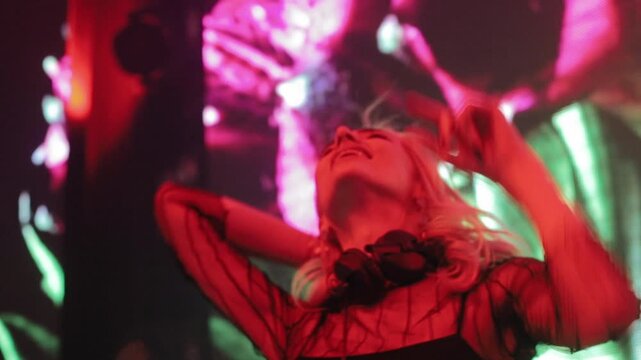 Smiling blonde woman indoors in frontal pose with arms raised and one hand near hip, lit by vivid pink, red, and green stage style glow, with woman, blonde, female, smile detail, showing portrait