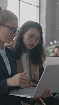 Two young businesswomen talking looking at laptop screen standing in busy office. Coworking and professional communication concept.