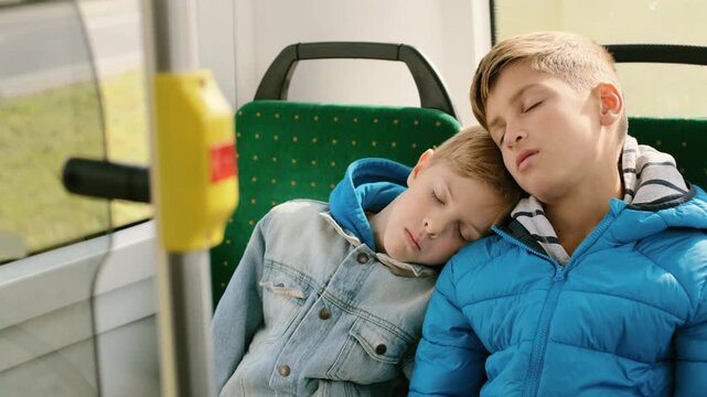 Two young boys asleep side-by-side on green patterned public transit seat, older boy on right wearing blue puffer jacket with striped hood lining and younger boy on left wearing light denim jacket