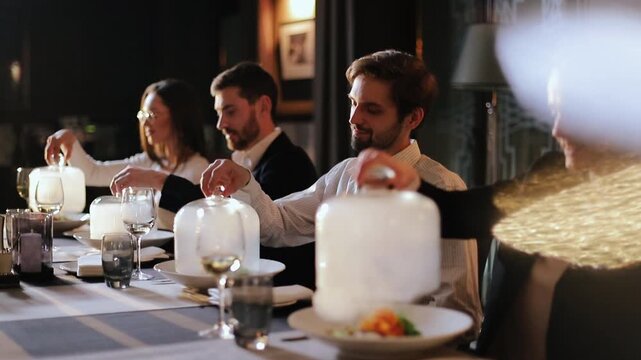 Group of adults seated along long table lift glass cloche lids in unison to reveal plated dishes beneath, visible steam or condensation inside cloches enhances reveal moment while hands hold glass