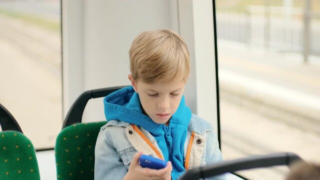 Close view of young boy riding daytime public transit, holding blue smartphone with both hands and looking down at screen with calm, concentrated expression inside clean urban bus interior