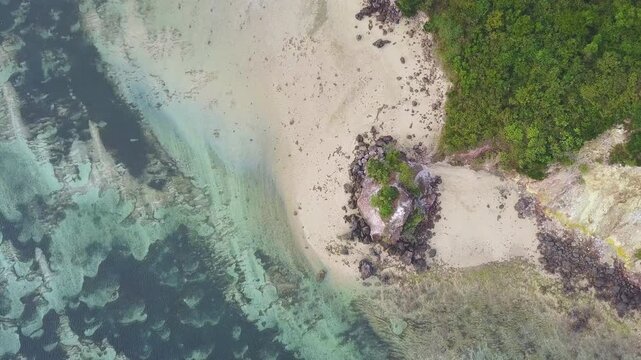 A drone descends and rotates over a rocky outcrop on a sandy beach with turquoise water in Lombok, Indonesia
