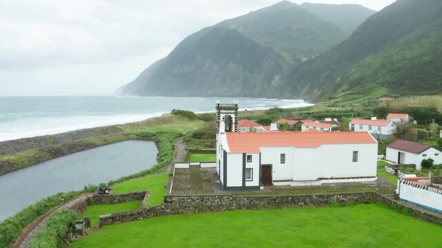 A white chapel with a red roof stands on green fields beside the lagoon at Faja de Santo Cristo on Sao Jorge with the surf behind.