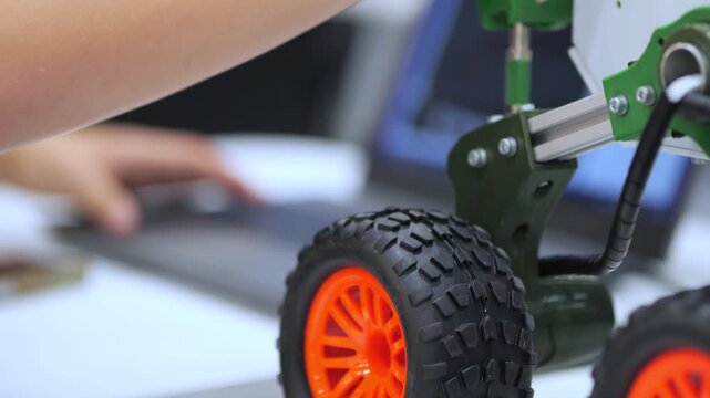 Close-up of child's hand adjusting small robot with orange wheels. Background shows blurred hands typing on laptop. Focus on mechanical details and bright colors. Suitable for STEM, education