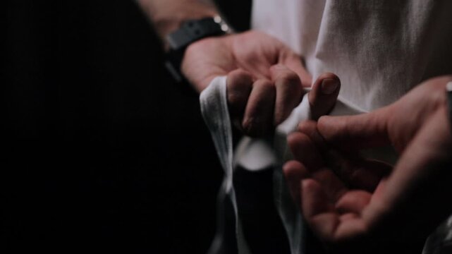 Hands and torso of adult shown close-up while tying white fabric strip around waist, fingers thread and pull cloth into knot with strands hanging downward, with hand, fabric, wrapping, bandage detail