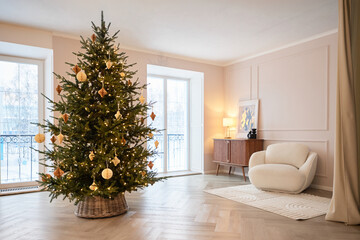 A Christmas tree with paper ornaments and glowing lights stands in a beige interior beside a cream armchair and wooden sideboard. Tall windows brighten the elegant holiday decor with winter daylight. © Yulia Mihaylova