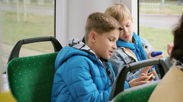 Two school-age boys seated side-by-side on public transit, both focused on handheld smartphones, foreground boy in bright blue puffer jacket and striped hood looks down at device with brief puckered
