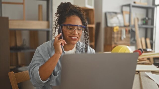 Woman carpenter at laptop holds smartphone to ear smiling while wearing goggles in studio; confidence planning collaboration.