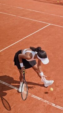 Vertical video of young Biracial woman serving tennis ball on clay court in slow motion from high angle. Ideal for sports apparel advertising and athletic social media campaigns