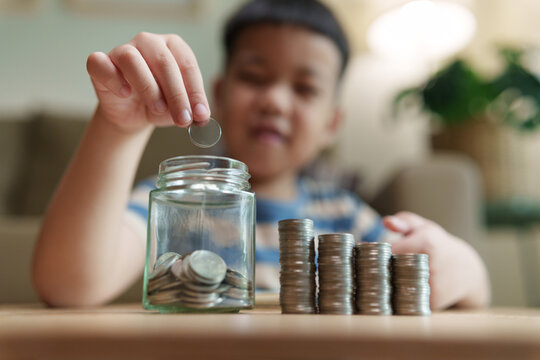 Asian boy putting a coin into a savings jar beside growing stacks of coins, representing financial education, money saving, budgeting, future planning, wealth growth and responsibility.