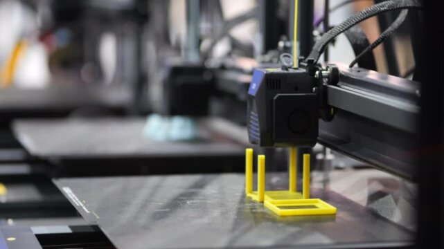 3D printer builds yellow object on metal surface. Machine moves precisely with cables and motors. Background shows blurred industrial equipment. Print head deposits material layer by layer