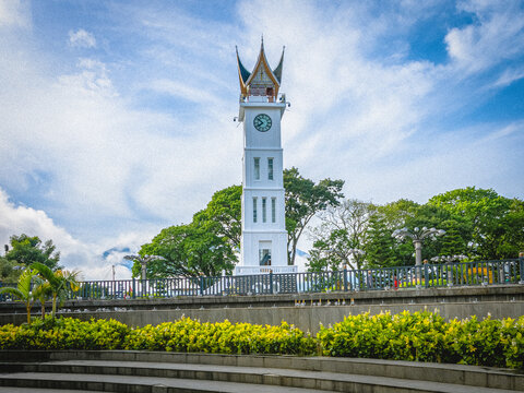 Bukittinggi, West Sumatra, Indonesia - September 21, 2026: Jam Gadang clock tower surrounded by trees and landscaped gardens under a partly cloudy blue sky.