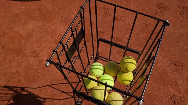 Caucasian young adult male player taking yellow tennis ball from basket hopper on clay court. Perfect for sports academy commercials and tennis coaching promotions