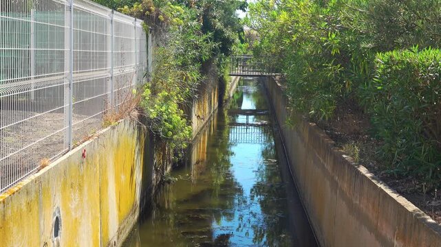 A waterway flows beside a fence and plants. Valras, France .