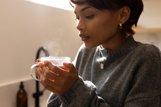 African American woman wearing grey knitted sweater holding steaming glass mug at kitchen sink