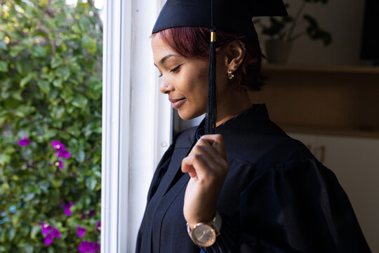 Woman standing by window at home holding mortarboard tassel while wearing graduation gown and watch