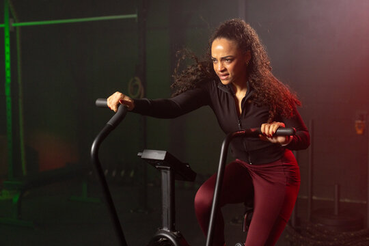 African American woman pedaling on air bike in gym, gripping handles with console, in athletic wear