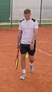Vertical video of CaucBiracial young adult man holding racket and bouncing ball while walking on clay tennis court in slow motion. Clip suits sports marketing, workout apps and athletic campaigns