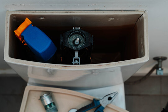 Cracked ceramic toilet cistern opened for repair with flush valve, pliers and a hose clamp on the removed lid. Home plumbing repair and water leak troubleshooting for sanitary ware replacement.