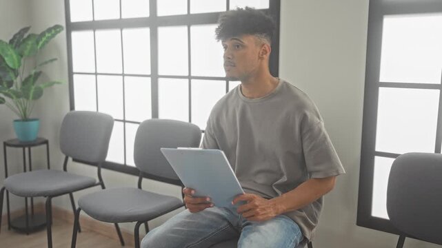 Man seated on a chair reading documents and checking wristwatch while holding a clipboard in a bright building waiting area; quiet anticipation.