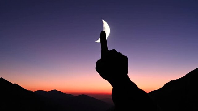 Silhouette of hand pointing at crescent moon over mountains