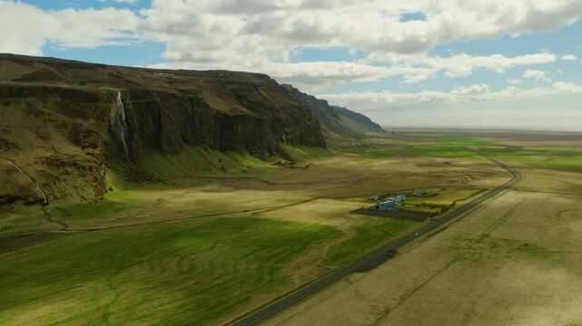 Foss a Sidu cascades from a basalt cliff above the farmstead and flat lowlands of Skaftarhreppur, Southern Region, Iceland, drone orbit.