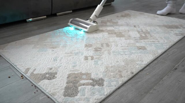 White cordless vacuum cleaner with turquoise LED illumination gliding across a patterned fabric rug to remove scattered particles from flooring