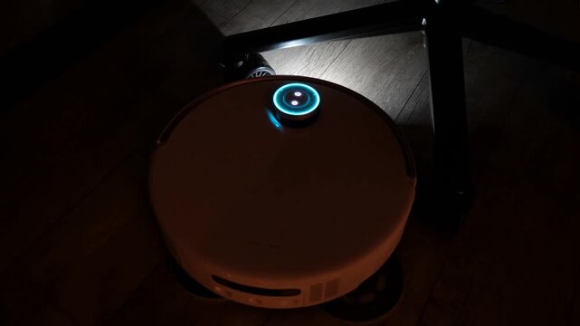 White automated floor cleaning machine navigating around furniture legs during evening hours in a modern domestic interior environment