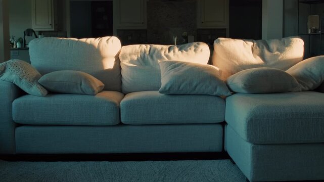 Wide slow zoom in shot of soft low-angle warm sunlight patterns hitting a beige sectional sofa with scattered cushions in a quiet living room, suggesting home comfort and relaxed lifestyle scenes.
