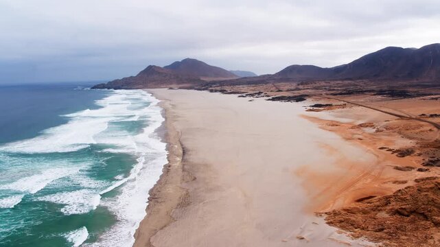 Turquoise waves crash along a vast, empty desert coastline where arid mountains meet the Pacific, sunlit patches of ochre sand adding warmth to this remote, untouched Atacama shoreline.