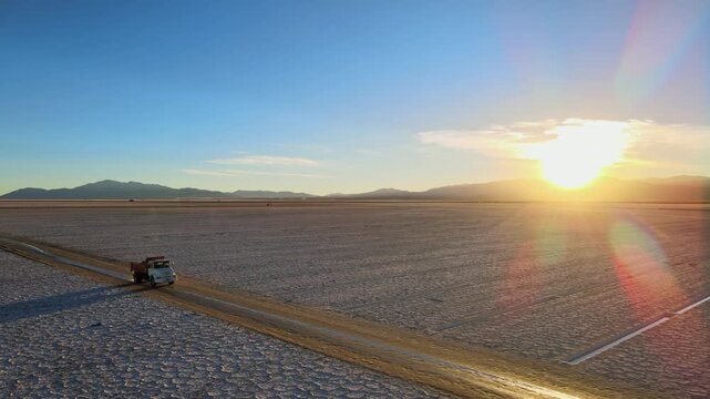 Utility truck drives along a muddy track across the white Salinas Grandes salt flat at sunrise, with mountains on the horizon and warm desert light.