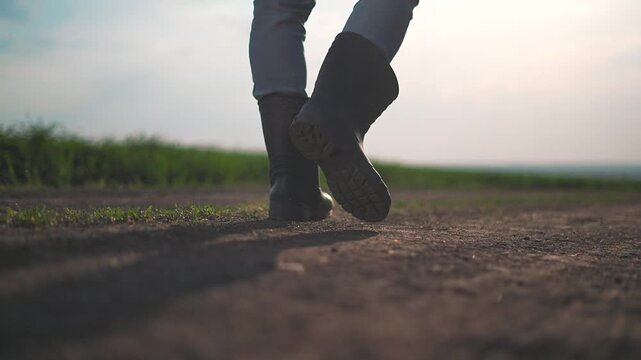 Field surrounds farmer walking along wide rural dirt path. Boots shift forward slow motion over soil. Outdoor landscape with field extends calmly while walking farmer moves through open natural space.