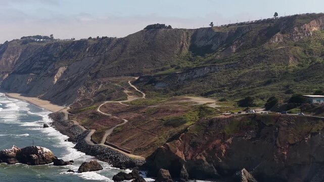 Majestic aerial footage capturing the rugged cliffs, sandy beach, and winding trails of Mussel Rock Park in Daly City, California, where the San Andreas Fault meets the Pacific Ocean