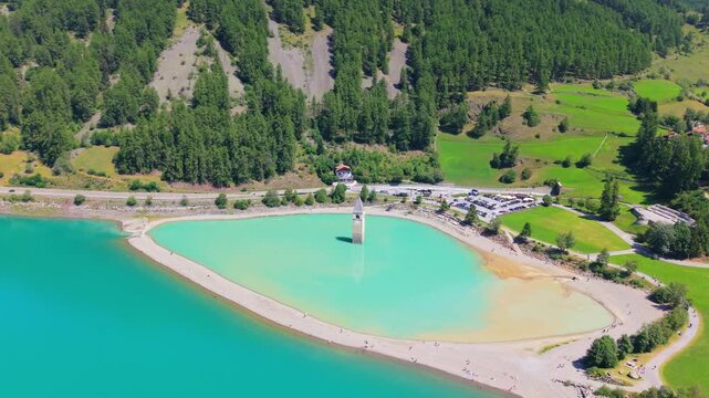 Aerial view of the iconic submerged church tower of Curon rising from the turquoise waters of Lake Resia, surrounded by alpine meadows and mountains in South Tyrol, Italy