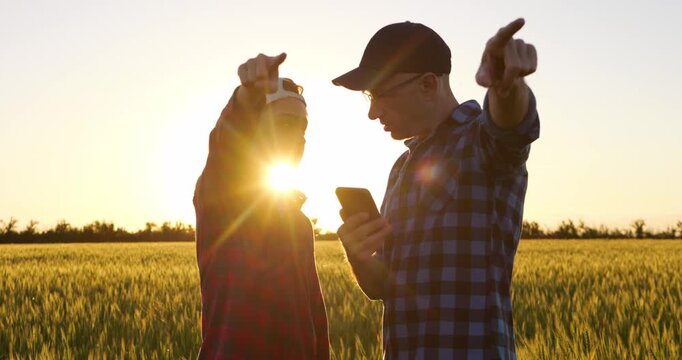 Father and son discuss farming plans in wheat field at sunset, using smartphone and pointing toward horizon. Countryside scene represents guidance, shared vision and future of family farming.